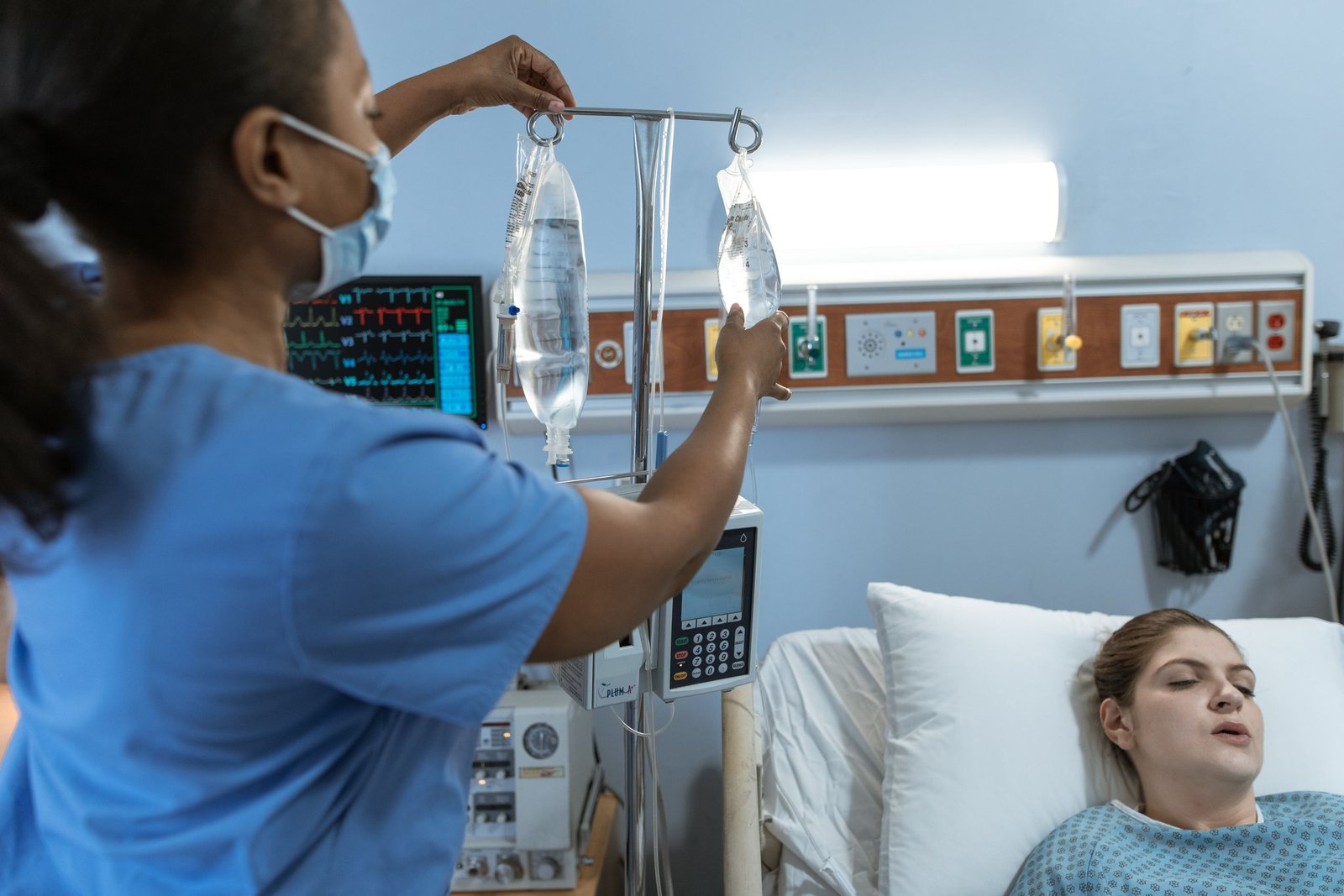 A nurse in a hospital setting adjusts an IV drip for a patient lying in bed, showcasing medical care.