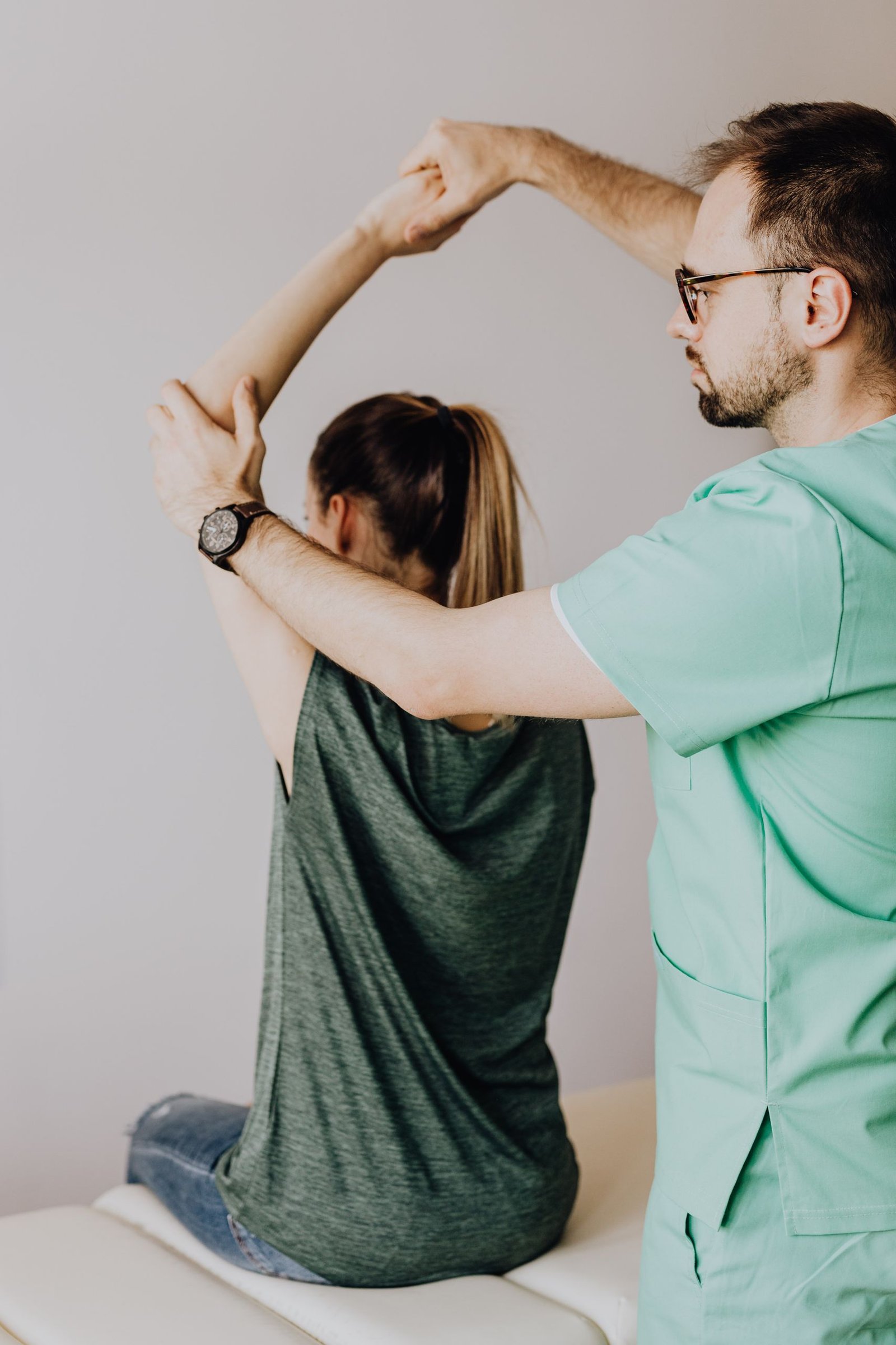 Crop osteopath in eyewear and uniform with wristwatch raising hand of unrecognizable slim woman sitting on examination couch in hospital